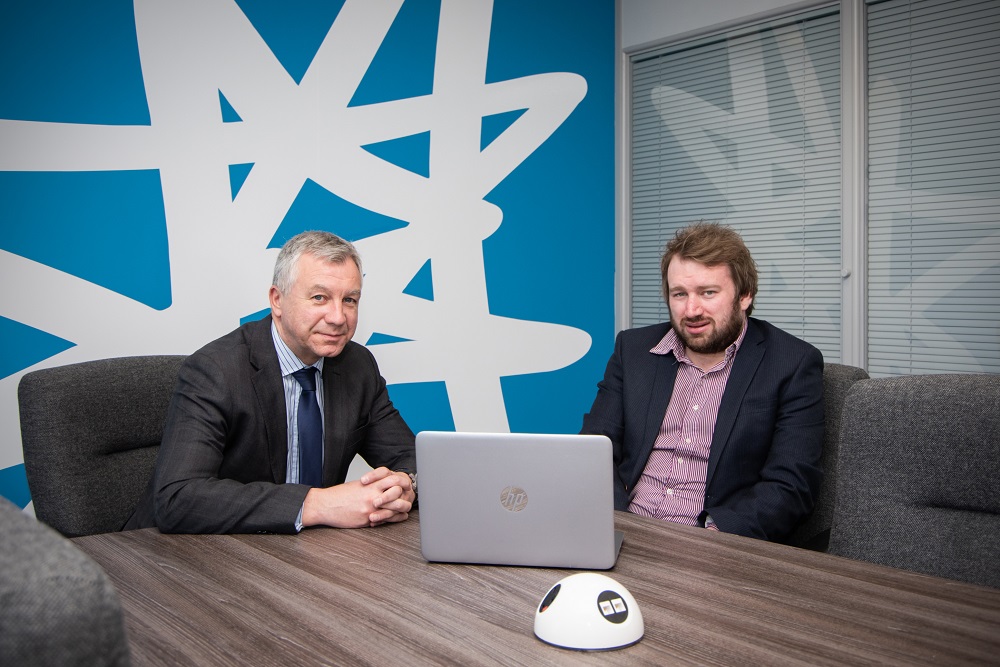 Two men in an office sitting behind a desk with a bright blue and white patterned wall behind a grey haired man.