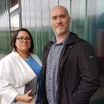 Man and woman outside green industrial building. Woman wearing glasses and has dark shoulder length hair to the left of the man is wearing a white jacket and a light blue top.