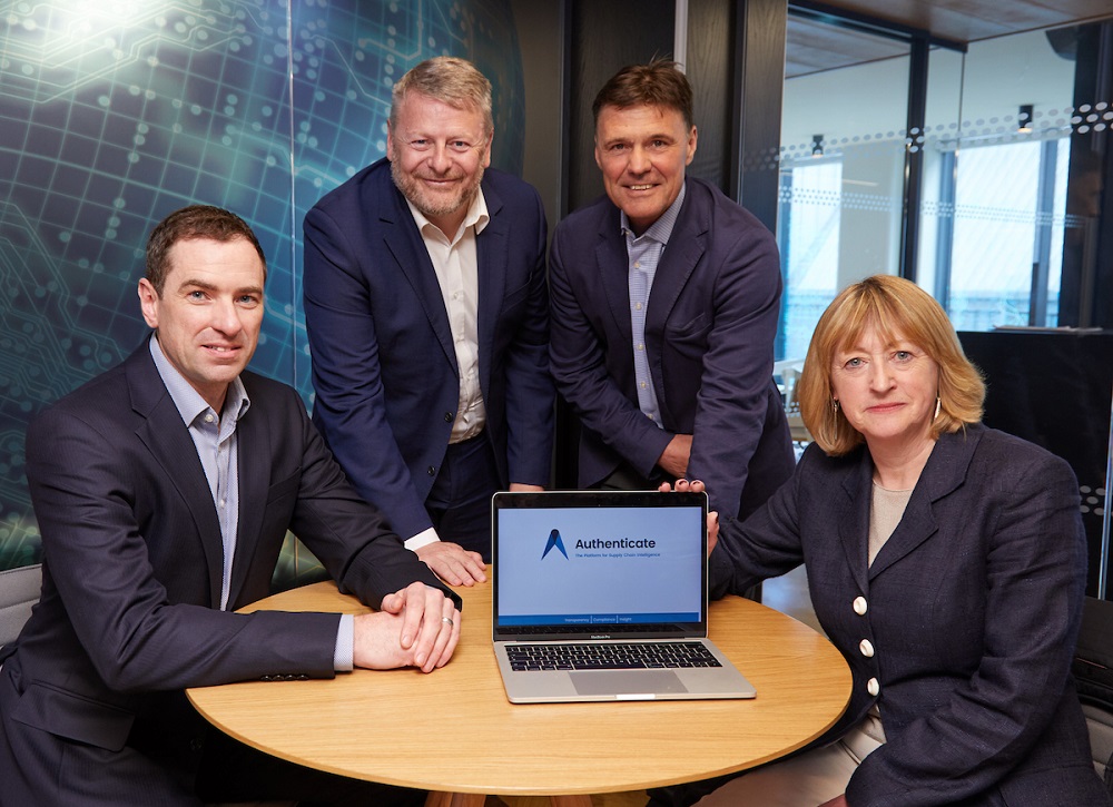 Four people around a table. There is only one woman and she is seated on the far right, the man to the left of the woman is wearing a dark blue suit and has dark hair, the man next to him has grey hair and the final man to the left is seated wearing a dark blue suit. The two men in the middle are standing up.