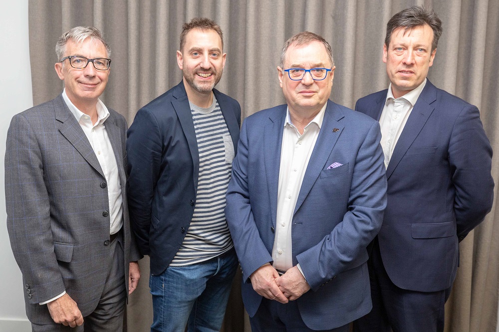 Four men standing up in front of a grey curtain. The man to the far right is wearing a blue suit and white shirt, next to him to his right is a man wearing blue rimmed glasses and to the far left is a man with grey hair and glasses wearing a dark grey suit.