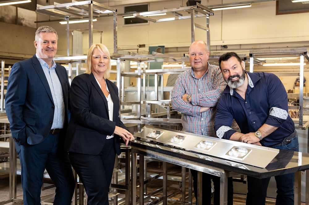 Three men and one blonde haired woman in a factory standing near machinery.