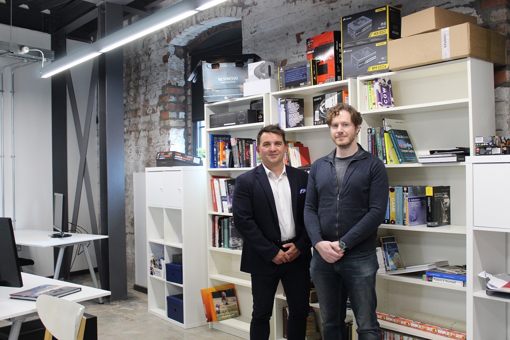 Two men in an office. Man to the right has a beard and is wearing casual clothes and the other man is wearing a suit.