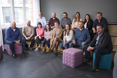 Large group of people in an office. There are two purple pouffes in the scene.