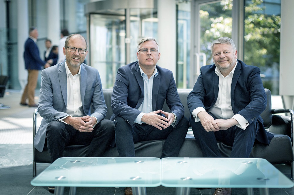 Three men sitting on a low leather sofa in an office. Man to the far right is wearing a dark blue suit, white shirt and is clasping his hands together, man in the middle is wearing glasses and is wearing a light blue shirt and man at the far left is wearing a light blue jacket and dark trousers.