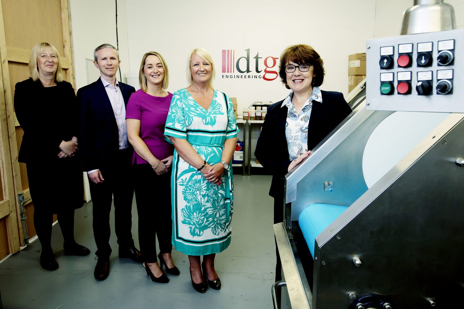 Five people in an industrial setting, woman to the far right wearing a black jacket and a patterned shirt is standing next to machinery and to the left of her is a blond haired lady wearing an acquamarine dress. There is only one man in the picture.