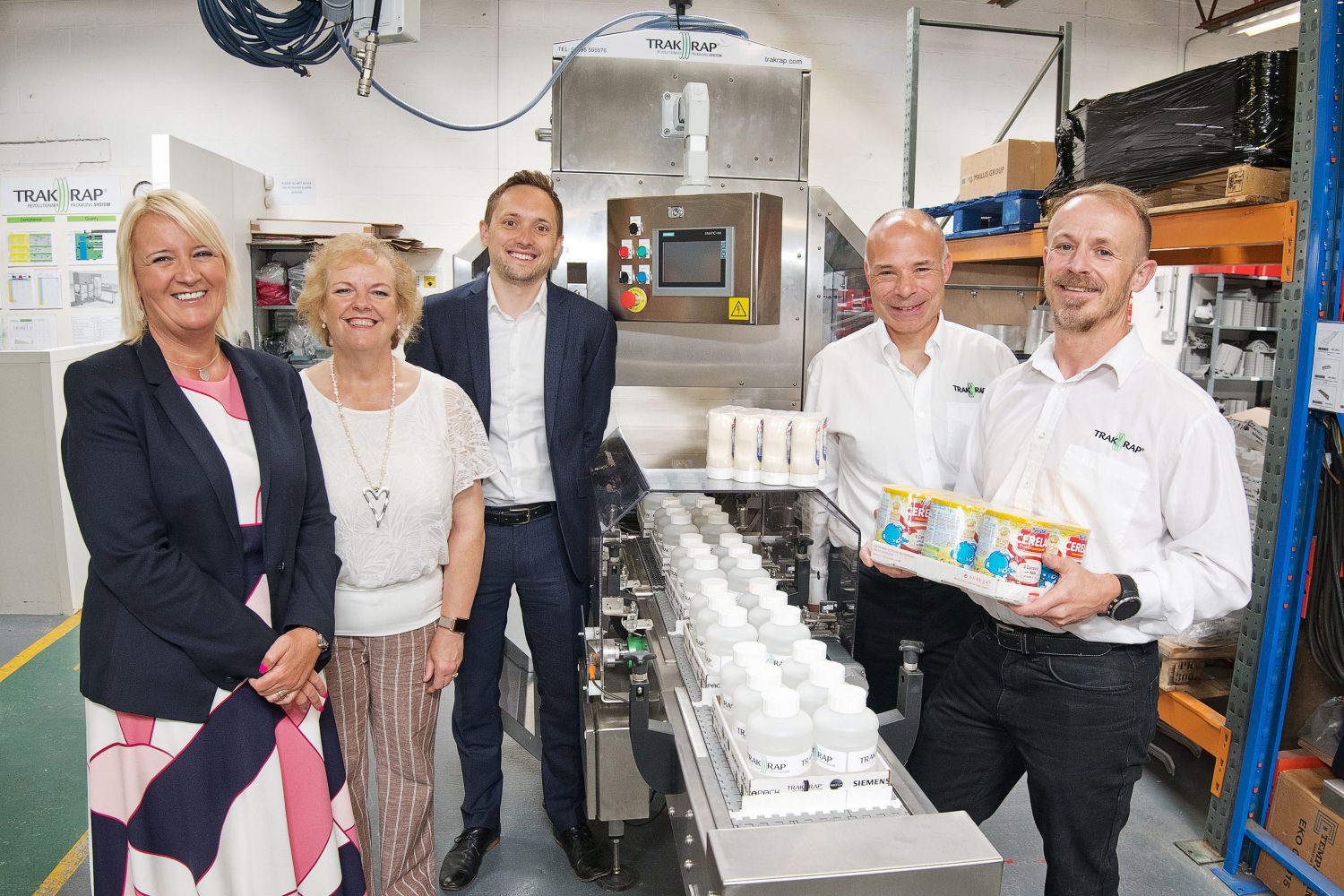 Three men and two women in a laboratory setting. Man to the far right is wearing a white shirt with Trak Rap branding and black trousers is holding some products, the man directly behind him is bald and is wearing a white shirt with Trak Rap branding on it. There is a blonde hair woman wearing a navy blue jacket and a pink, blue and white patterned dress on the left and behind her there is another woman with short curly hair wearing a white top and brown and white striped trousers. Finally there is a man behind the last woman wearing a navy blue suit and a white shirt.