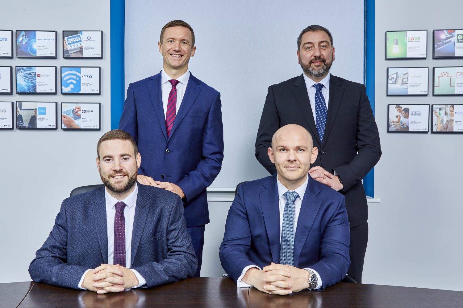 Four men in an office standing in front of pictures. There are two men seated in front a wooden table in front. One of them to the left has a beard and is wearing a blue jacket and the other man is bald. The remaining two men at the back are standing up. The man standing to the far right has a beard and the other man has a blue jacket, white shirt and red and blue striped tie.