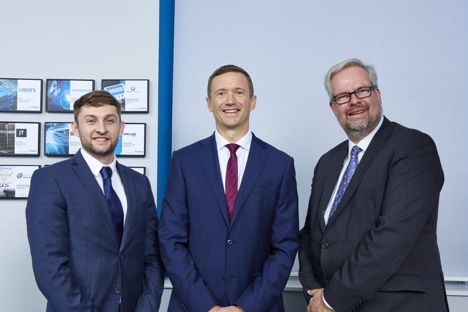 Three men in an office standing in front of pictures. Man to the far right has grey hair, glasses and a beard, he is wearing a navy jacket, man in the middle is wearing a blue jacket, red and blue striped tie and a white shirt and the man to the far left is wearing a blue jacket, white shirt and blue tie.