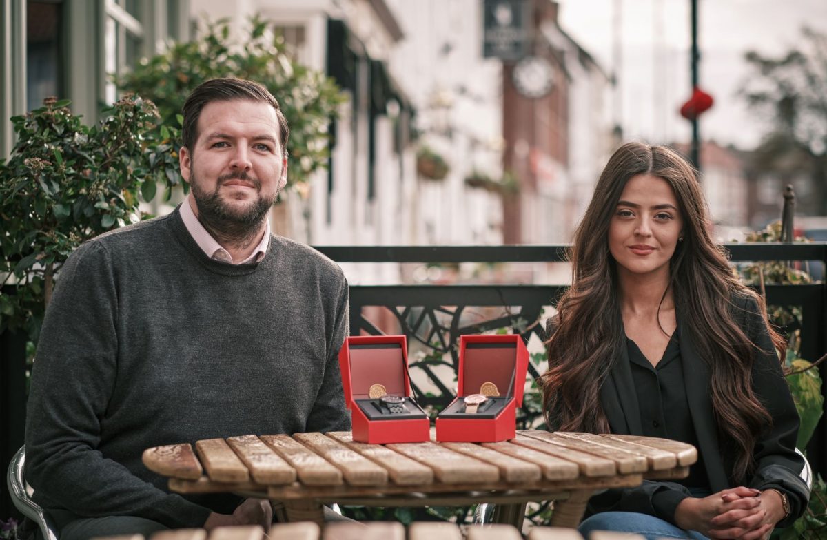 Young lady with long dark hair sitting to the right and bearded man to the left wearing a brown jumper. They are both sitting next to a table outside with two watches in boxes placed on a table.