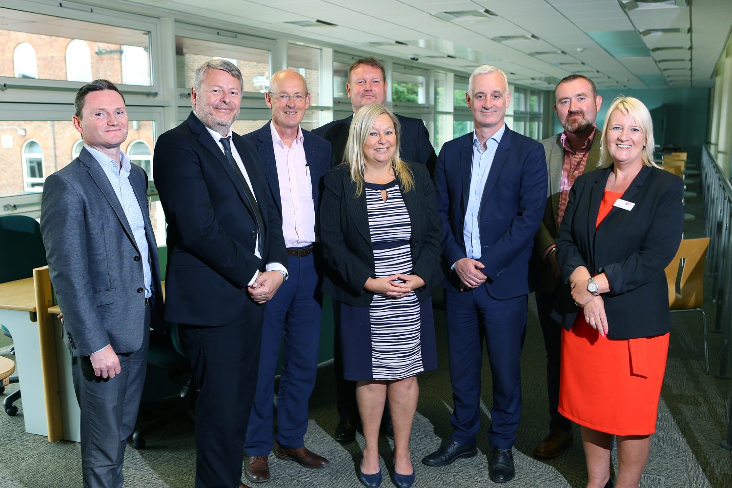 Eight people in an office. Woman to the far left has short blonde hair and is wearing a red dress and a black jacket, there is one other woman wearing a black and white stripey dress and has long blond hair. The man to the far left has a gunmetal grey suit on and to his right is a grey beared man wearing a black suit with a black tie.