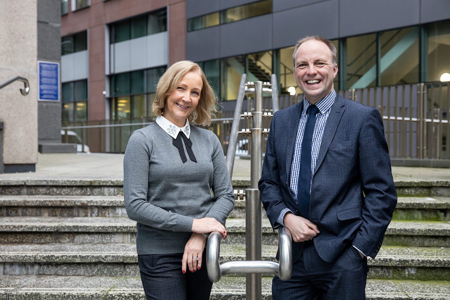 A woman outside an office building positioned to the left. She has a hand on a hand railing and has blonde short hair. She is wearing a grey jumper with a black bow on it and a pair of navy trousers. To the far right there is a man wearing a navy blue suit, blue tie and checked blue and white shirt. He too is leaning his arm on the handrail. Both people are shown in front of a flight of steps.