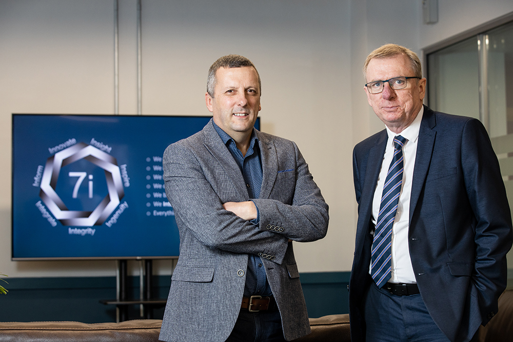 Two men standing in front of 7i poster in an office. Man to the far right is wearing glasses, has severely pockmarked skin and is wearing a blue jacket, white shirt and blue stripey tie.