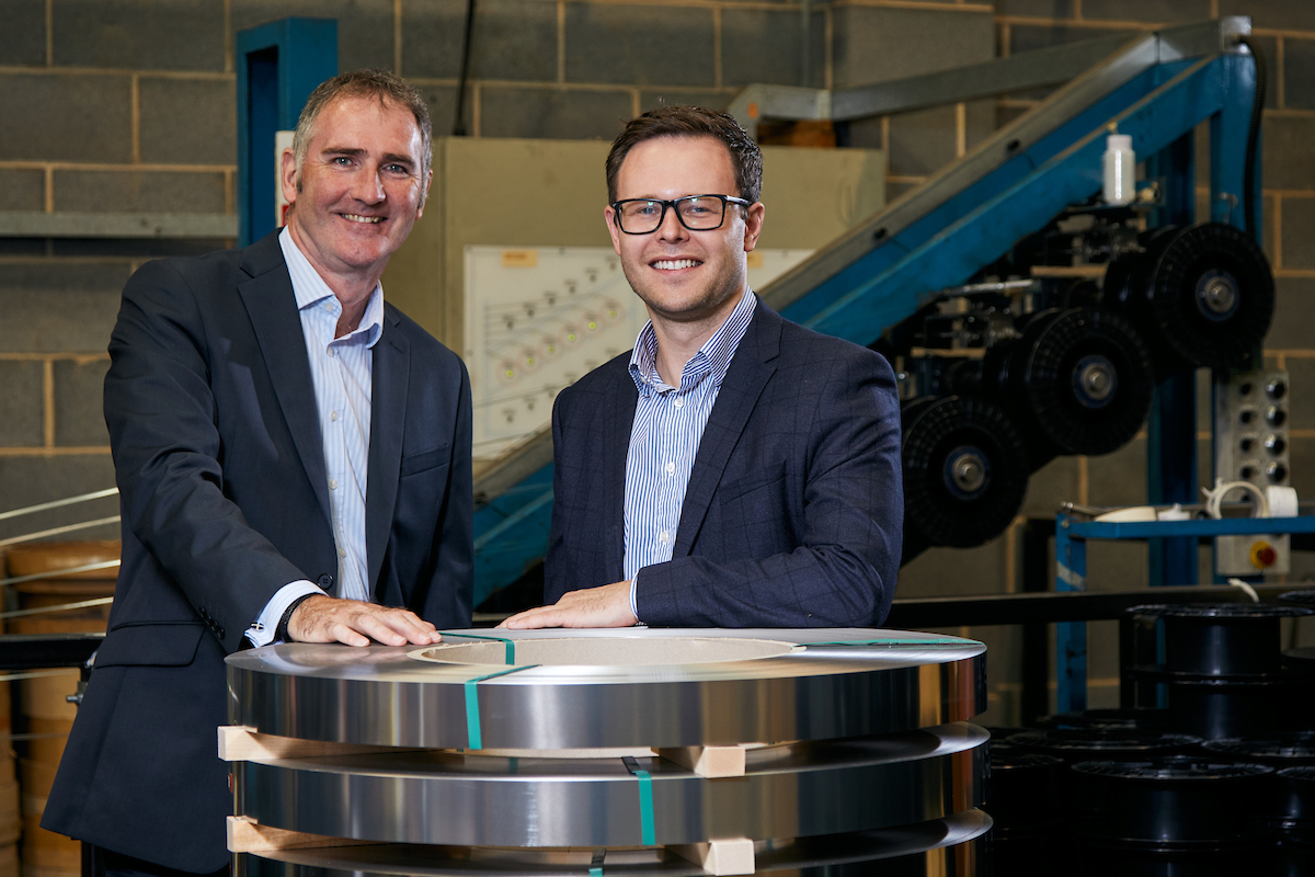 Two men standing in an industrial environment. Man to the far left has her hand on an industrial disc and is wearing a blue suit and a light blue shirt and the man to the right is wearing a blue jacket, white and blue stripey shirt and has dark hair and is wearing glasses.