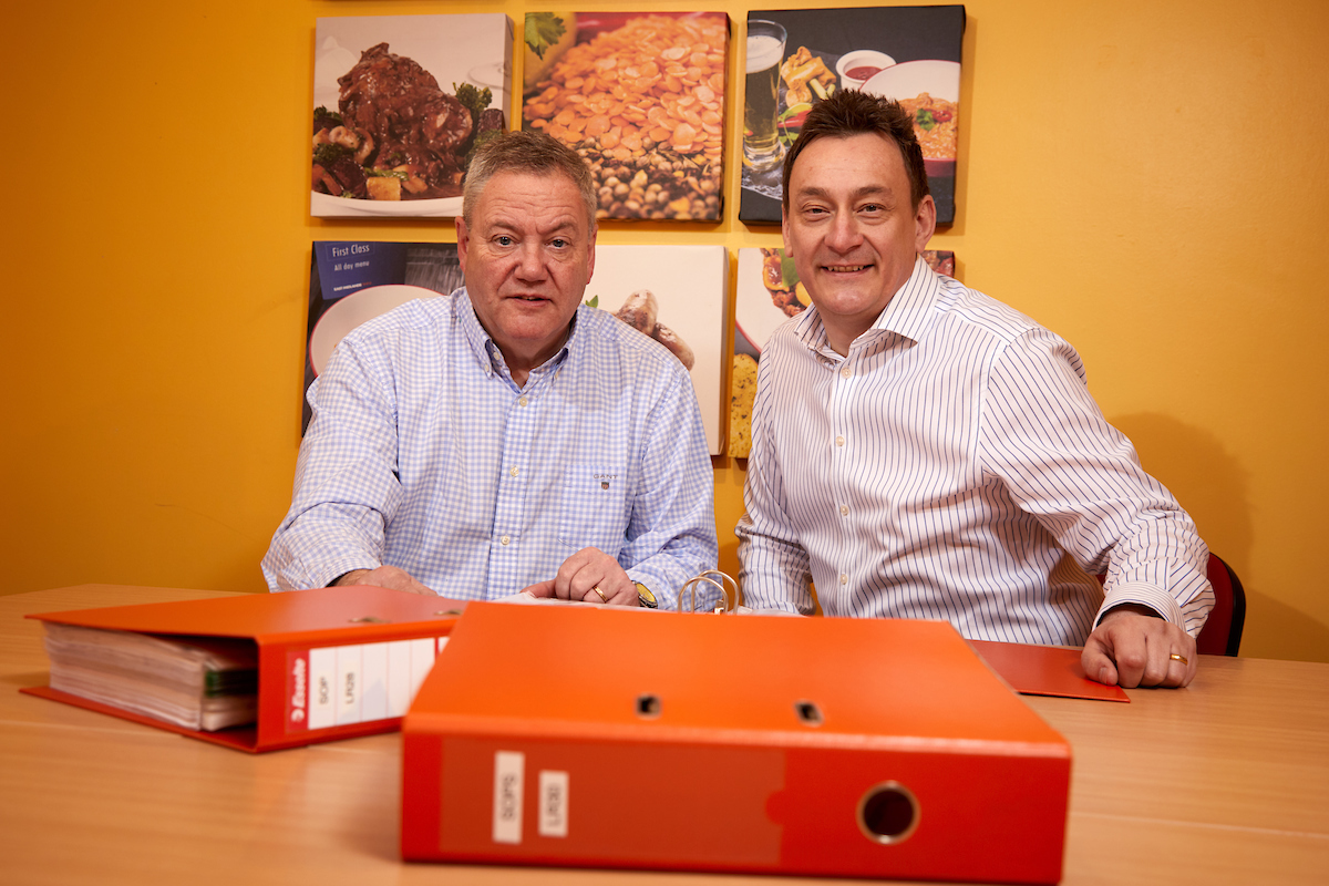 Two seated men in front of a poster of food against a yellow wall. There are two red folders on the desk. Man to the far right has dark hair and a white and blue stripey shirt.