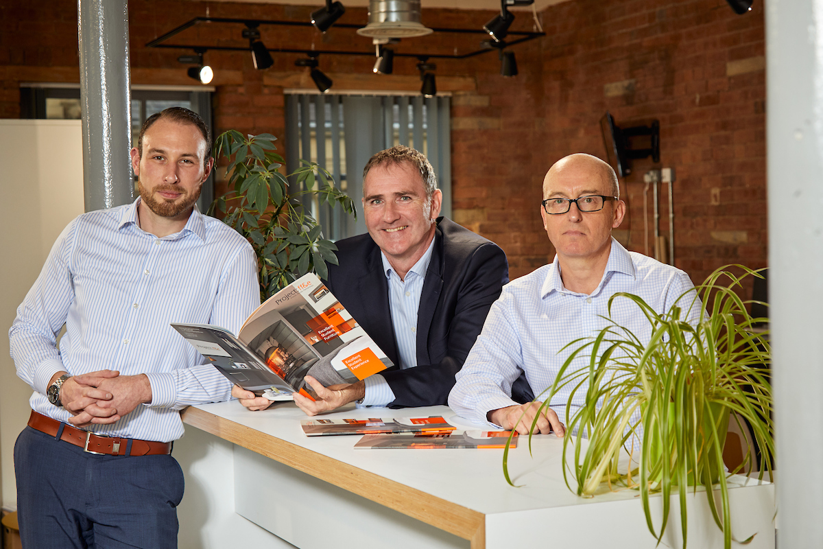 Three men standing around an office table. Two men are behind the table and the one to the far right is bald and has glasses. He is waring a light blue shirt and has a potted plant to the right of him. The other man behind the table is holding a brochure and is wearing a black jacket and light blue shirt. The remaining man is to the front of the table and is beared. He is wearing a pair of casual pair of blue trousers and a blue and white striped shirt.