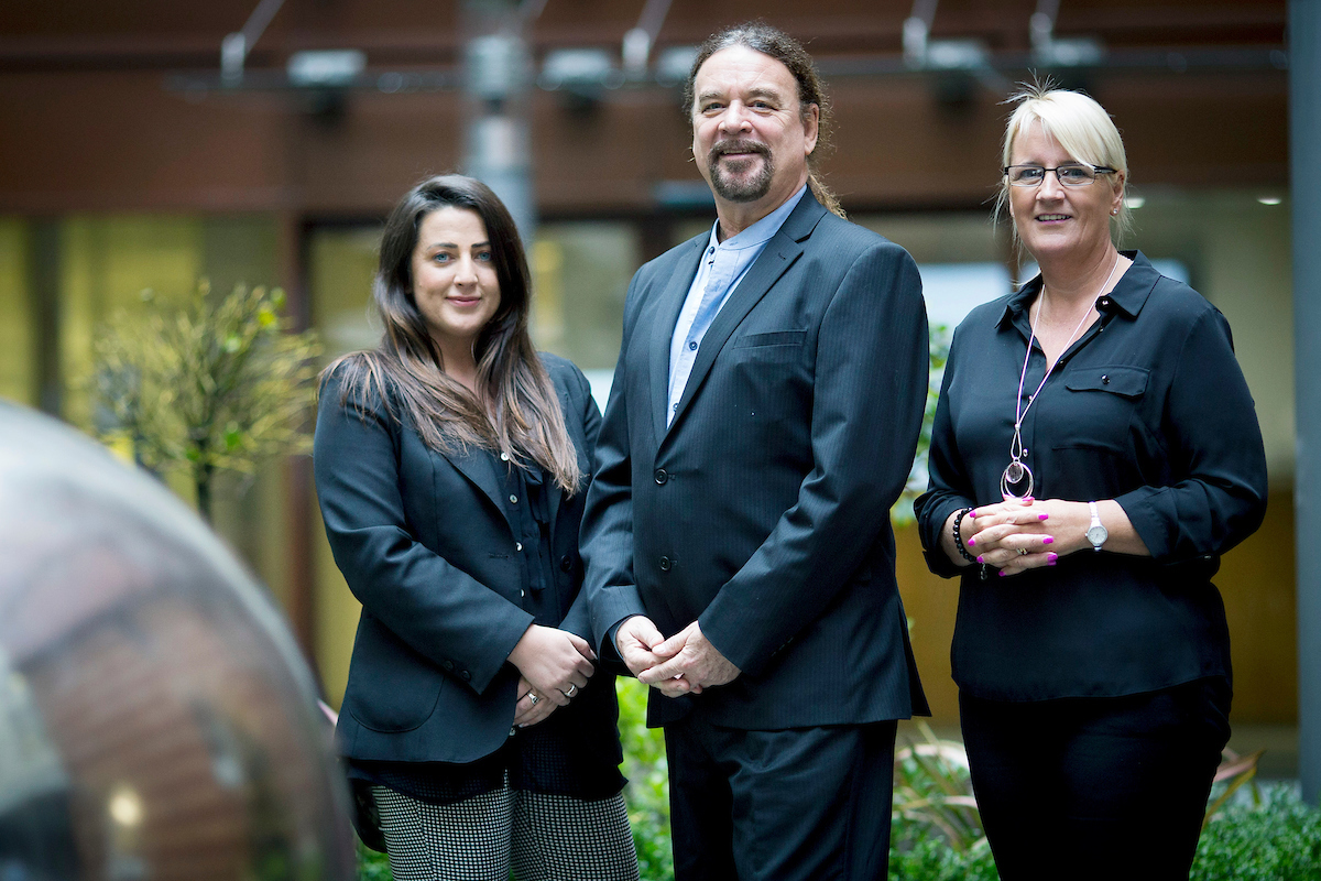 Three people outside, woman to the far right has blond hair and is wearing glasses. She is wearing a black suit and has a large necklace on, man in the middle has long dark hair in a pony tail and a beard. The woman on the far left has long dark hair and is wearing a black jacket and black and white checked trousers.