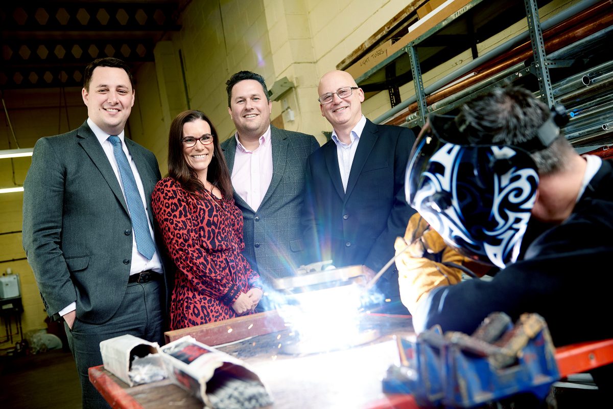 Four people in a line looking at a man welding wearing a head protection. The four people include one woman with glasses wearing a pink and black patterned dress. She is standing to the right of a man wearing a suit and blue tie.