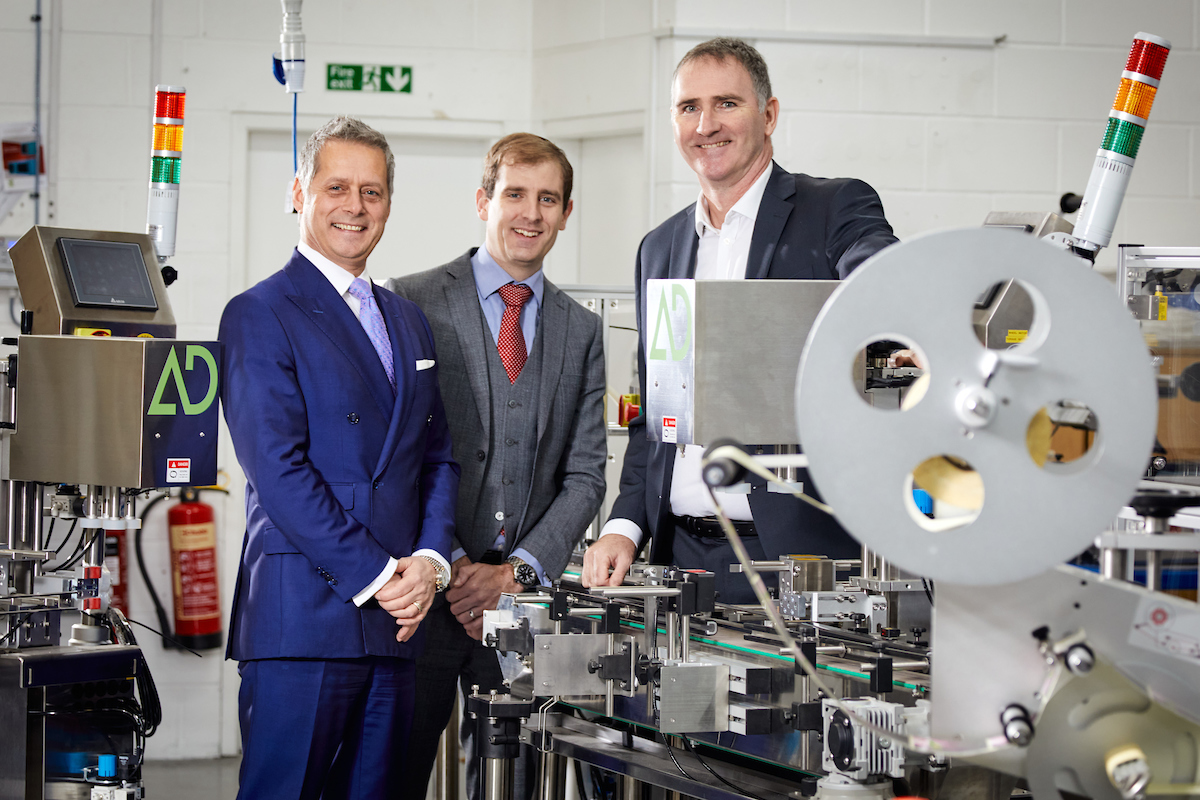 Three men in a manufacturing plant. Tall man in the far left is wearing a navy jacket and white shirt, man in the middle has a grey suit on with a matching waistcoat, blue shirt and red tie. The man to the far left is wearing a bright blue suit, white shirt and light blue tie.