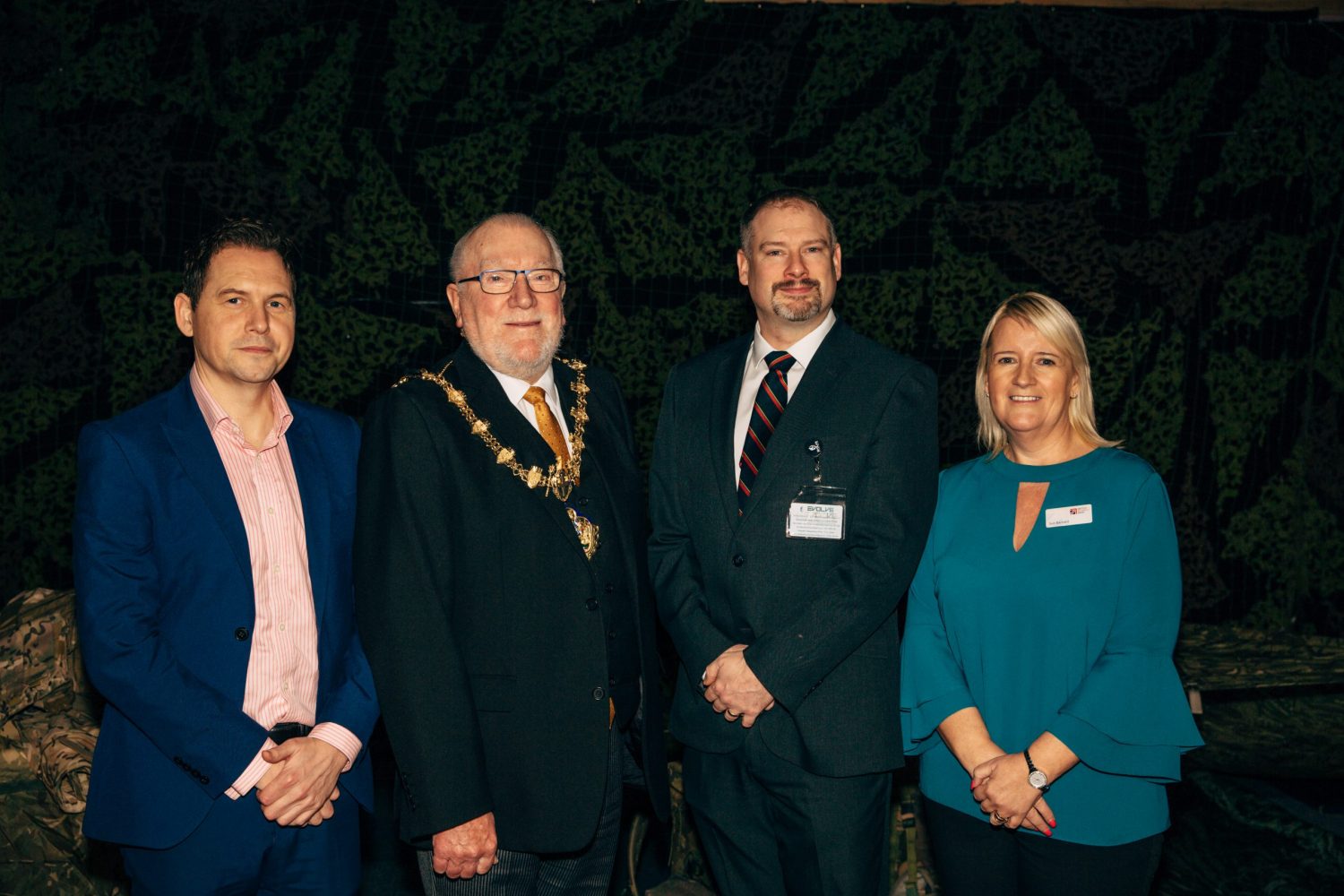 Four people at night, one woman in the group to the far right wearing a teal coloured top has short blonde hair and man to the far left has a blue suit with a coral shirt. There is an elderly man in the middle wearing mayoral regalia.