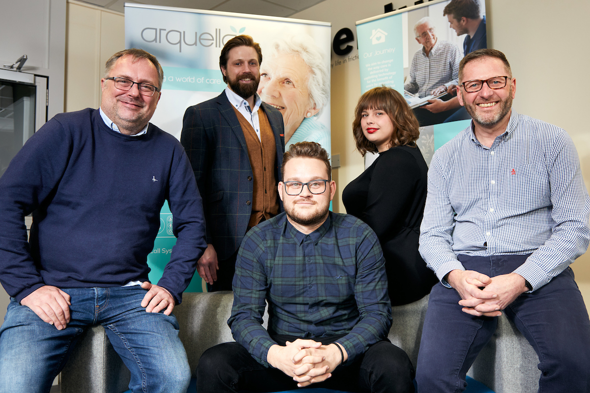 Five people in a group which includes one woman wearing a black jumper wearing red lipstick seating next to a bearded man to her left wearing a blue stripped jacket, brown cardigan