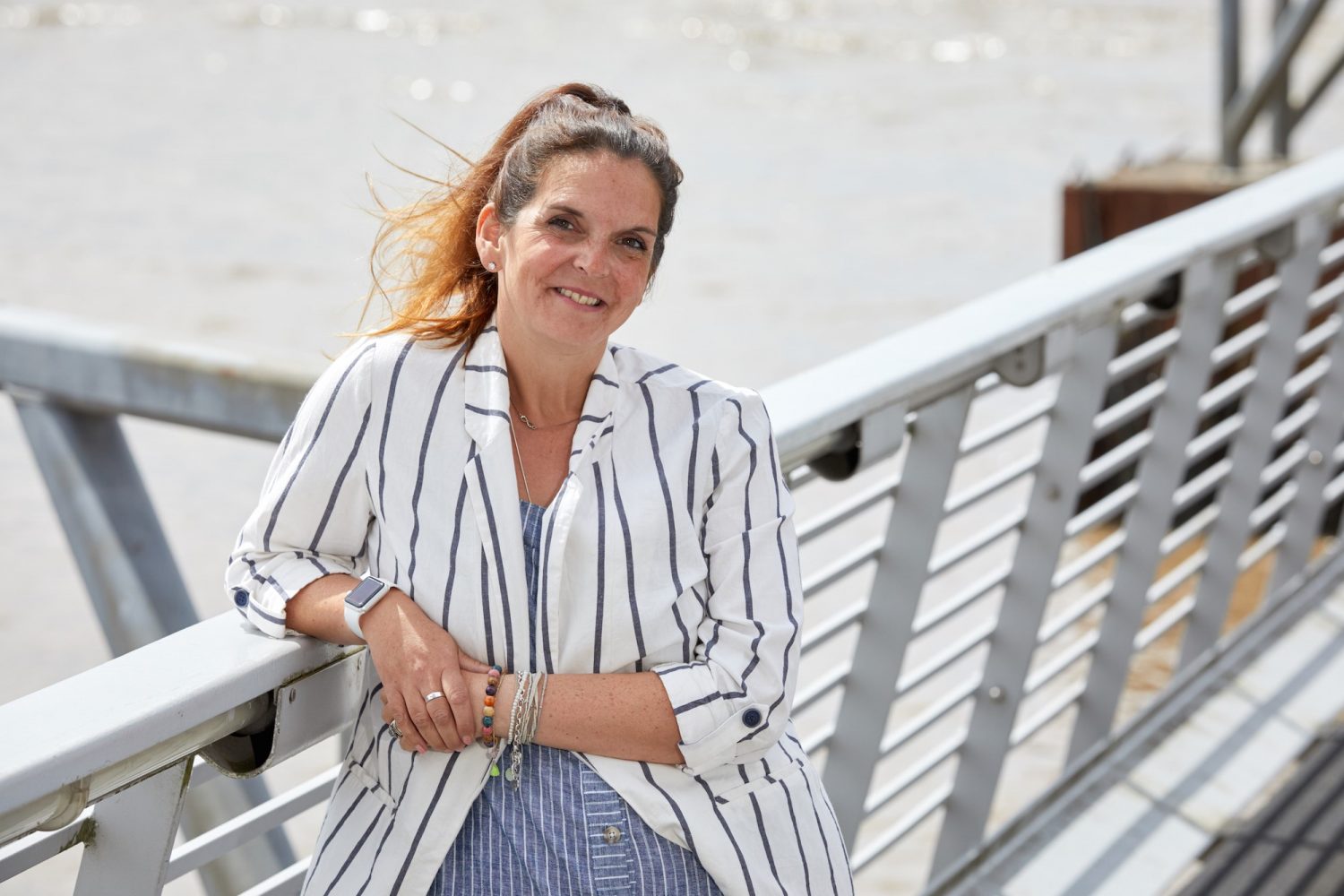 Woman leaning against white railings wearing a white and blue stripped jacket and blue and white stripped skirt. She has long hair tied back and has lots of bracelets on her right arm.