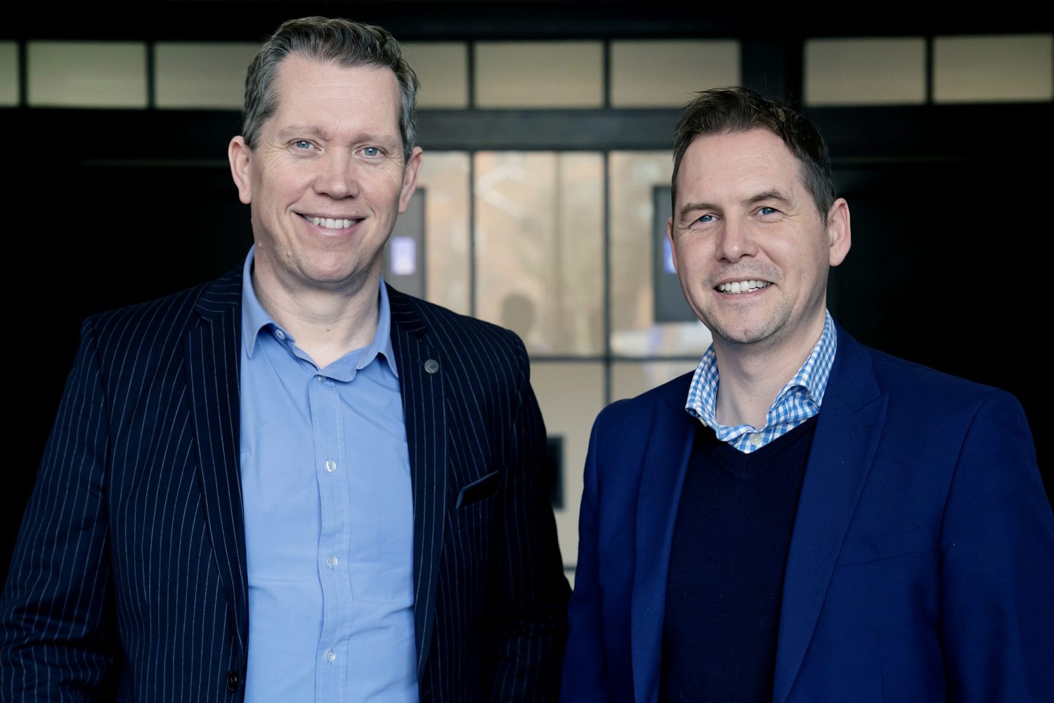 Two men standing in front of glass window. Man to the left wearing blue shirt and pinstripped jacket and man to the right wearing blue jacket, navy/black jumper and blue checked shirt.