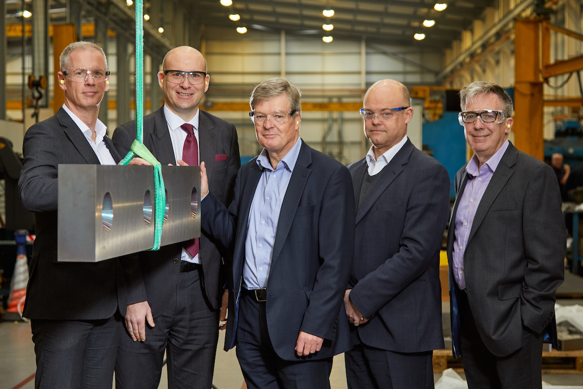 Five men inside a factory, they are all wearing protective glasses. Man to the far right is wearing a dark grey suit and a purple shirt and man to the far left has his hand on a rectangular piece of metal.