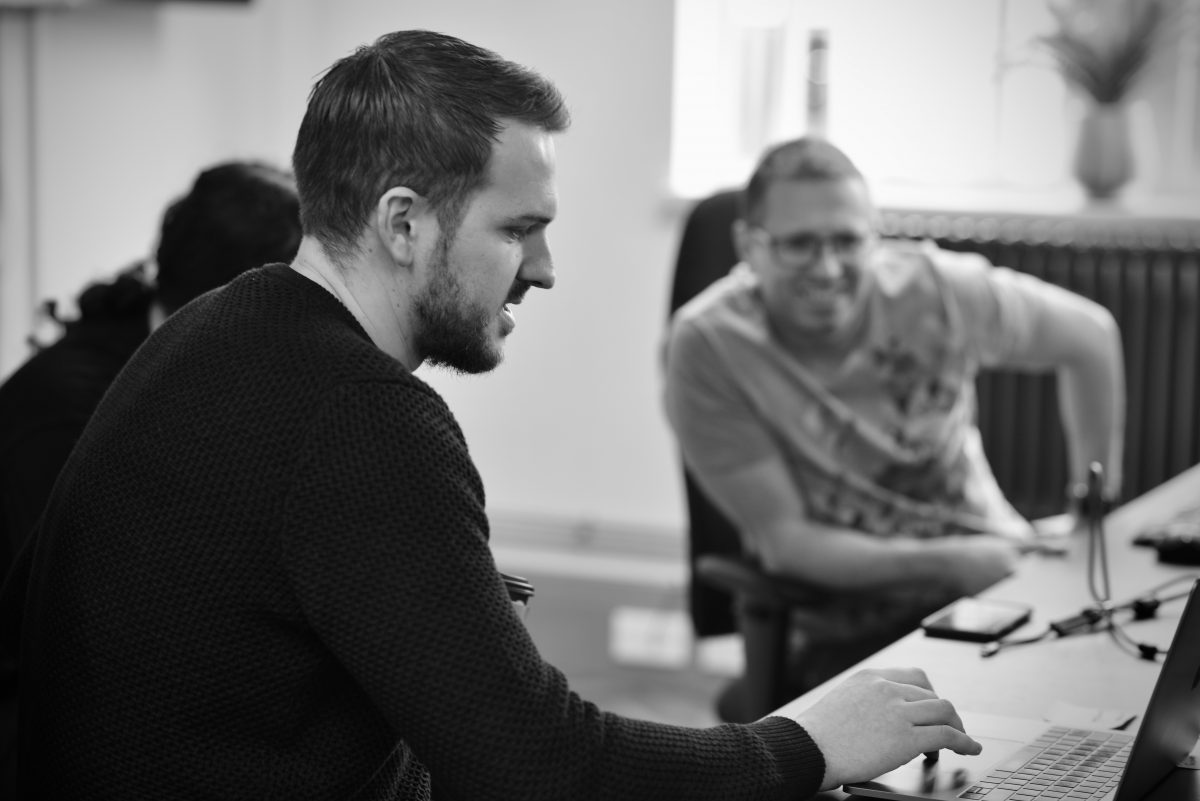 Mono image of two men in office environment. Man to left working on laptop.