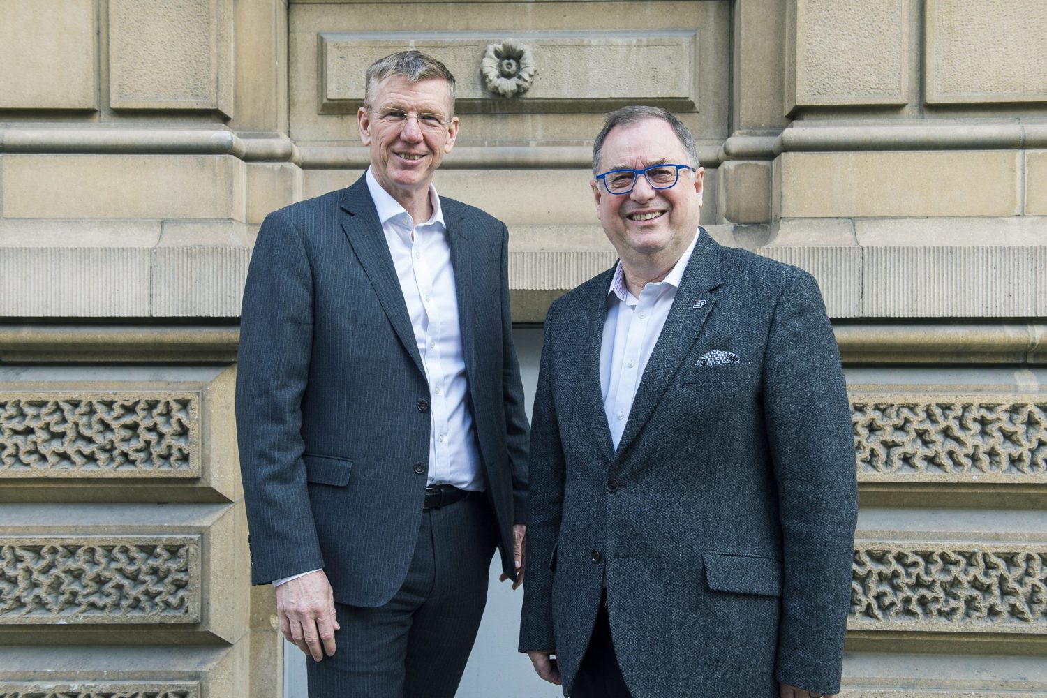 2 men stood in suits in front of a brick building